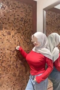 Woman in red top and hijab posing by tiled wall and mirror, reflecting her image.