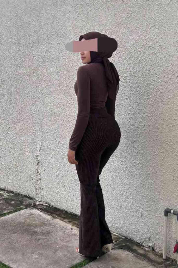 Woman in brown hijab and outfit standing against a textured wall.