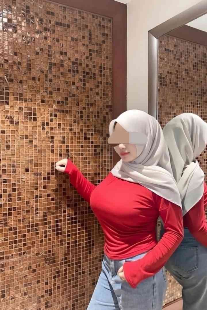 Woman in hijab, red top, and jeans posing against a textured wall, reflected in a mirror.