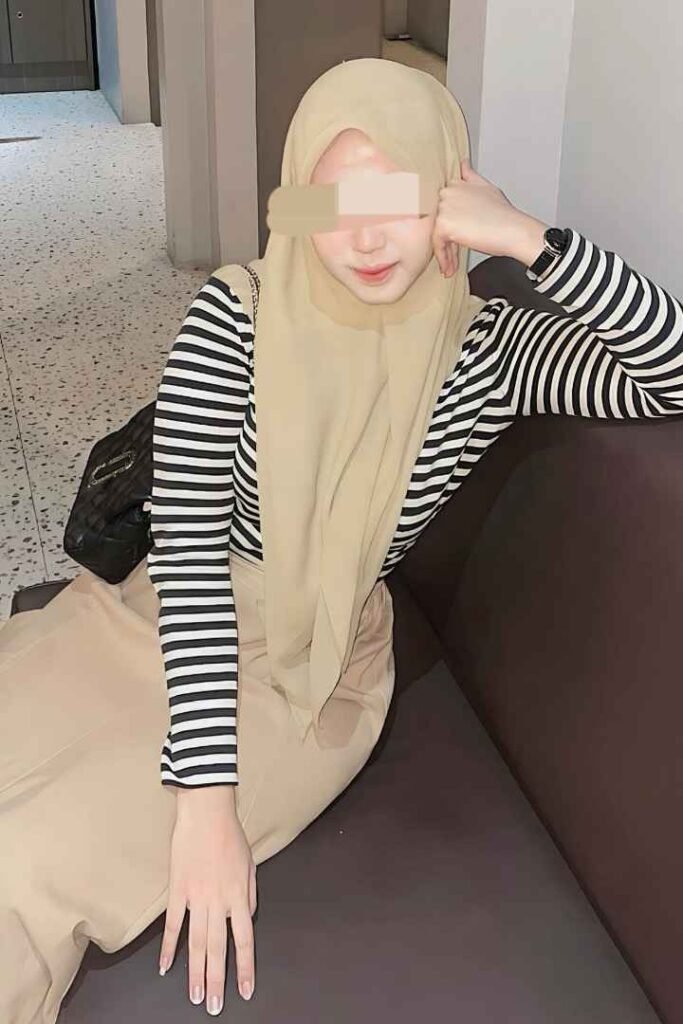 Woman in hijab, striped top, and beige pants sitting on a couch.