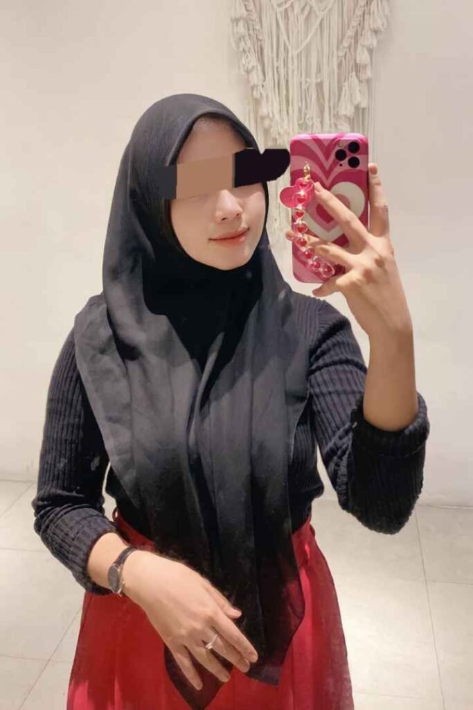 Woman in hijab with pink phone case, wearing a red skirt and black top.